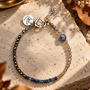 Handgefertigtes Edelsteinarmband mit facettierten dunkelmetallischen, rauchgrauen und tiefblauen Edelsteinen sowie silberfarbenem Verschluss und Anhänger. Das Armband liegt auf einem hellen, dezent strukturierten Untergrund und wird von warmem, natürlichem Herbstlicht mit sanften Schatten beleuchtet.