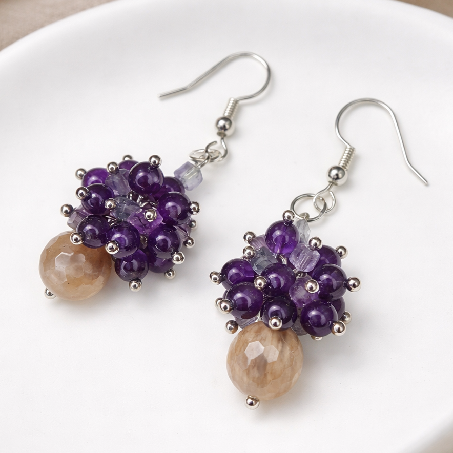 Zwei handgemachte Ohrringe aus Mondstein, Amethyst und Fluorit vor hellem Hintergrund mit weichem Schatten.