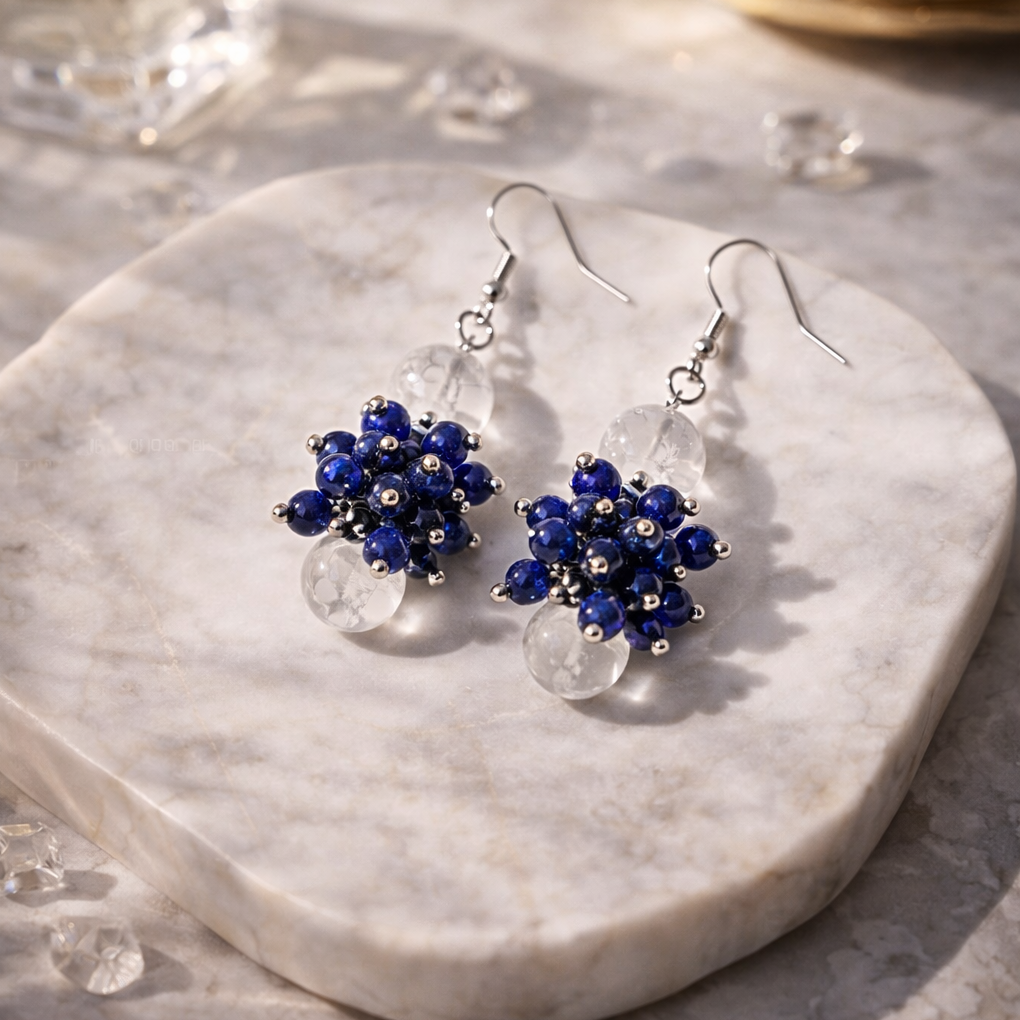 Luxuriöse Edelstein-Ohrringe „Blue Hour“ mit Lapislazuli, Bergkristall und Hämatit auf hellem Marmor, Soft-Fokus Hintergrund.