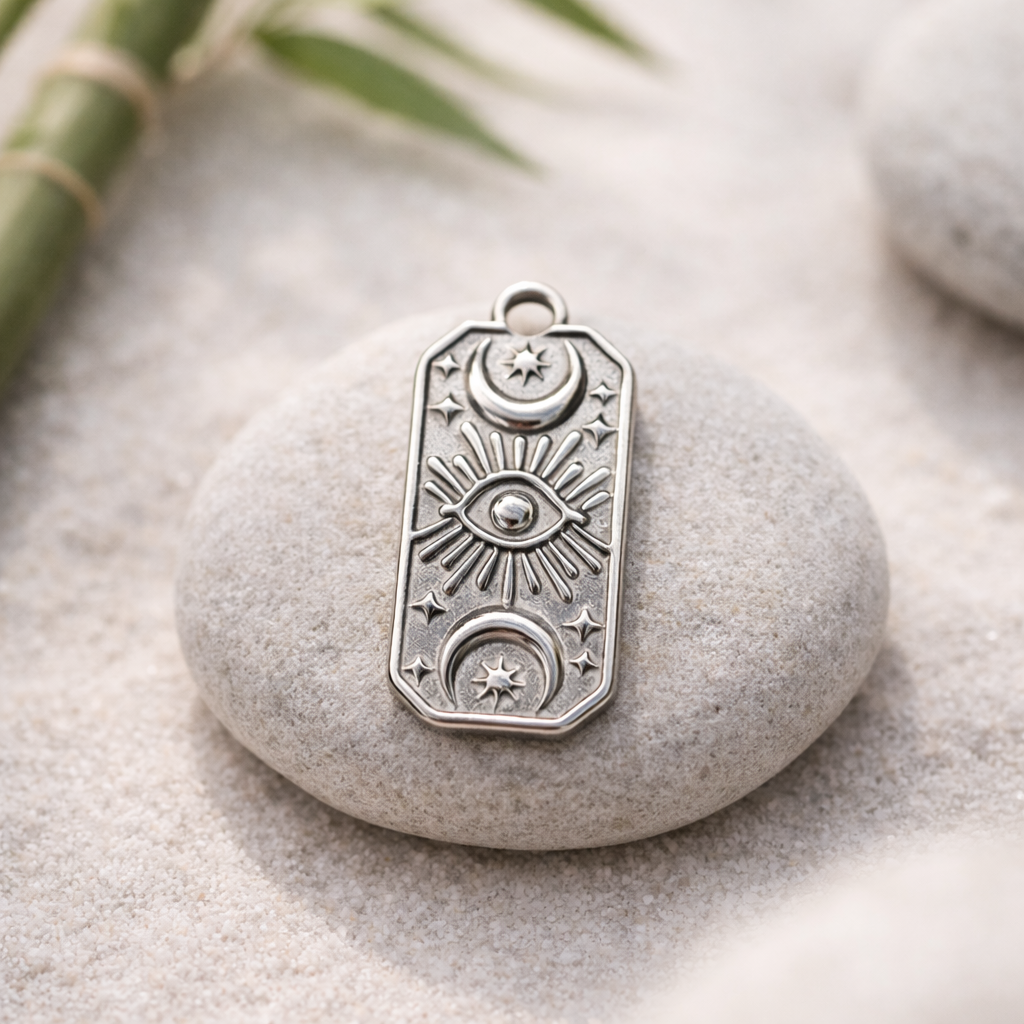 Mystic Eye Edelstahl-Charm ohne Kette auf glattem Flussstein in feinem Sand, Zen-Natur-Stillleben mit weichem Tageslicht und unscharfem Bambus im Hintergrund