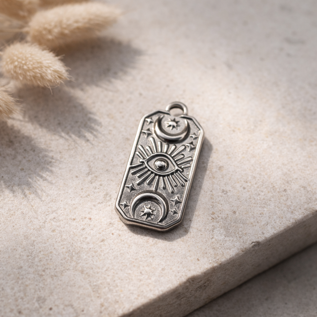 Mystic Eye Edelstahl-Charm ohne Kette auf glattem Flussstein in feinem Sand, Zen-Natur-Stillleben mit weichem Tageslicht und unscharfem Bambus im Hintergrund